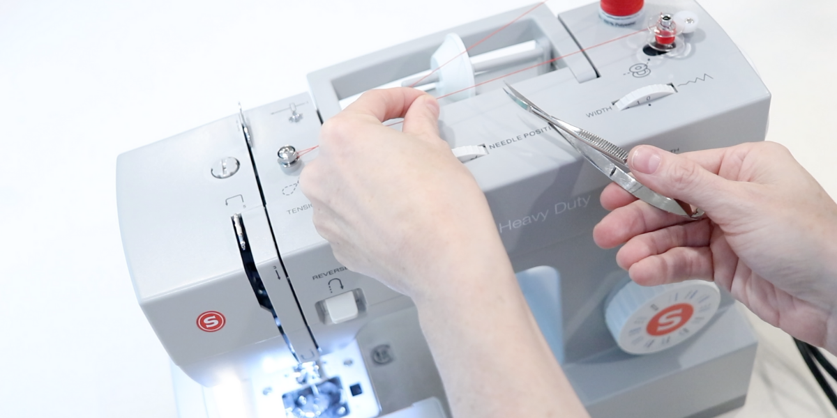 a girls hand using small scissors to clip the red thread on a sewing machine. Blog post about how to thread a sewing machine